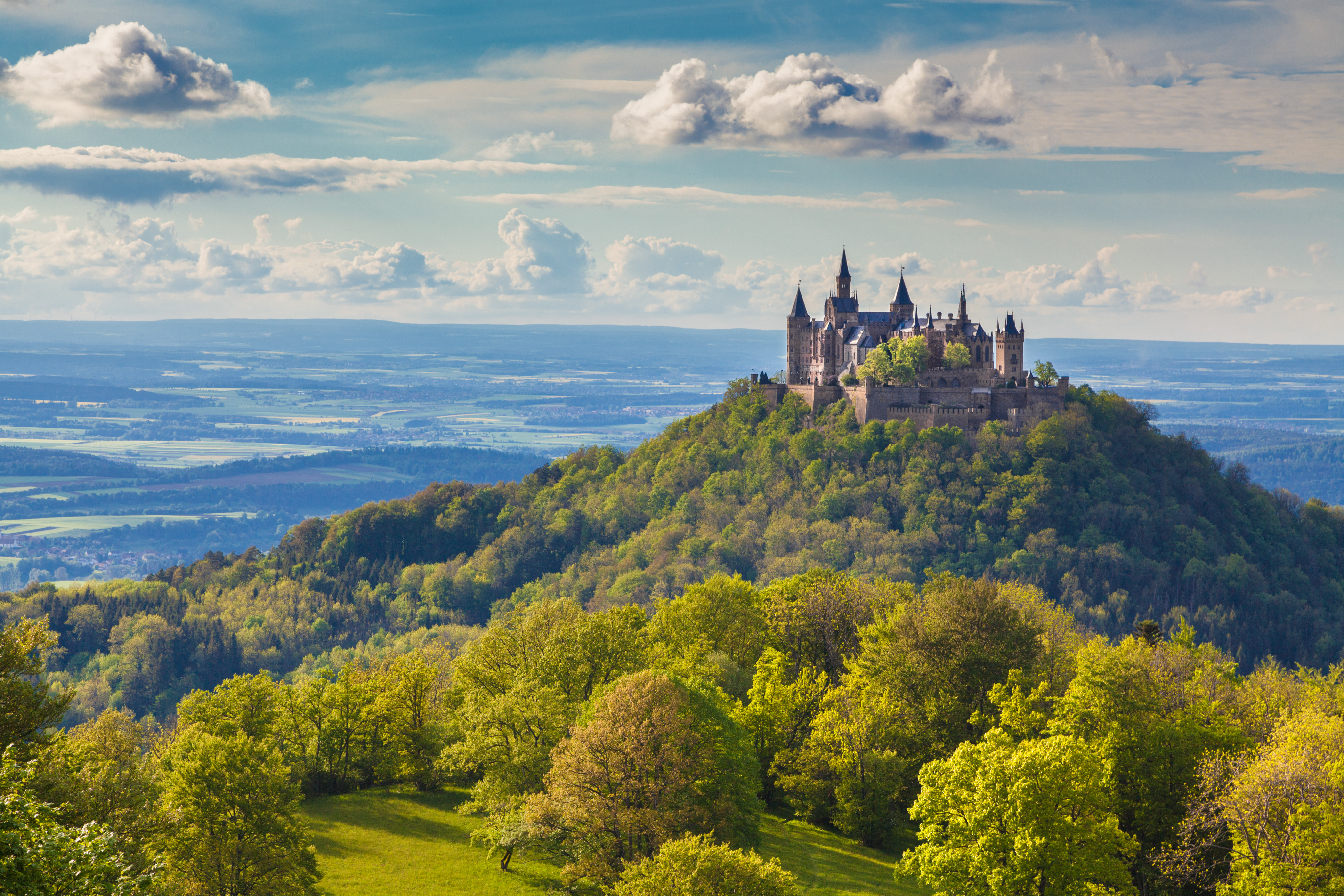 Das Bild zeigt eine Fotoaufnahme der Burg Hohenzollern auf der schwäbischen Alb. Die Aufnahme ist vom Zeller Horn in der Nähe von Onstmettingen aufgenommen. Die Burg trohnt auf einem Hügel zwischen grünen Wäldern und Wiesen. Im Hintergrund ist der Zollernalbkreis und ein strahlend blauer Himmel zu sehen. Das Bild zeigt eine Fotoaufnahme der Burg Hohenzollern auf der schwäbischen Alb. Die Aufnahme ist vom Zeller Horn in der Nähe von Onstmettingen aufgenommen. Die Burg trohnt auf einem Hügel zwischen grünen Wäldern und Wiesen. Im Hintergrund ist der Zollernalbkreis und ein strahlend blauer Himmel zu sehen.